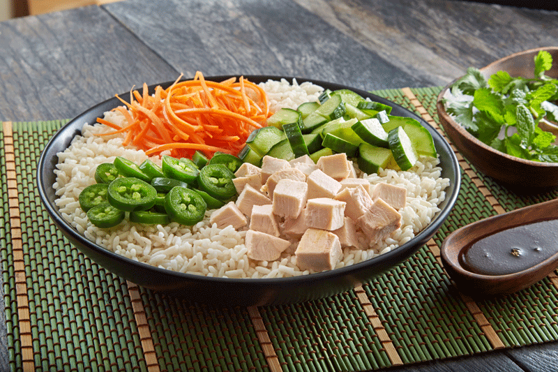 Shredded carrot, cucumber, pepper and chicken with sauce to the side over a bowl of rice