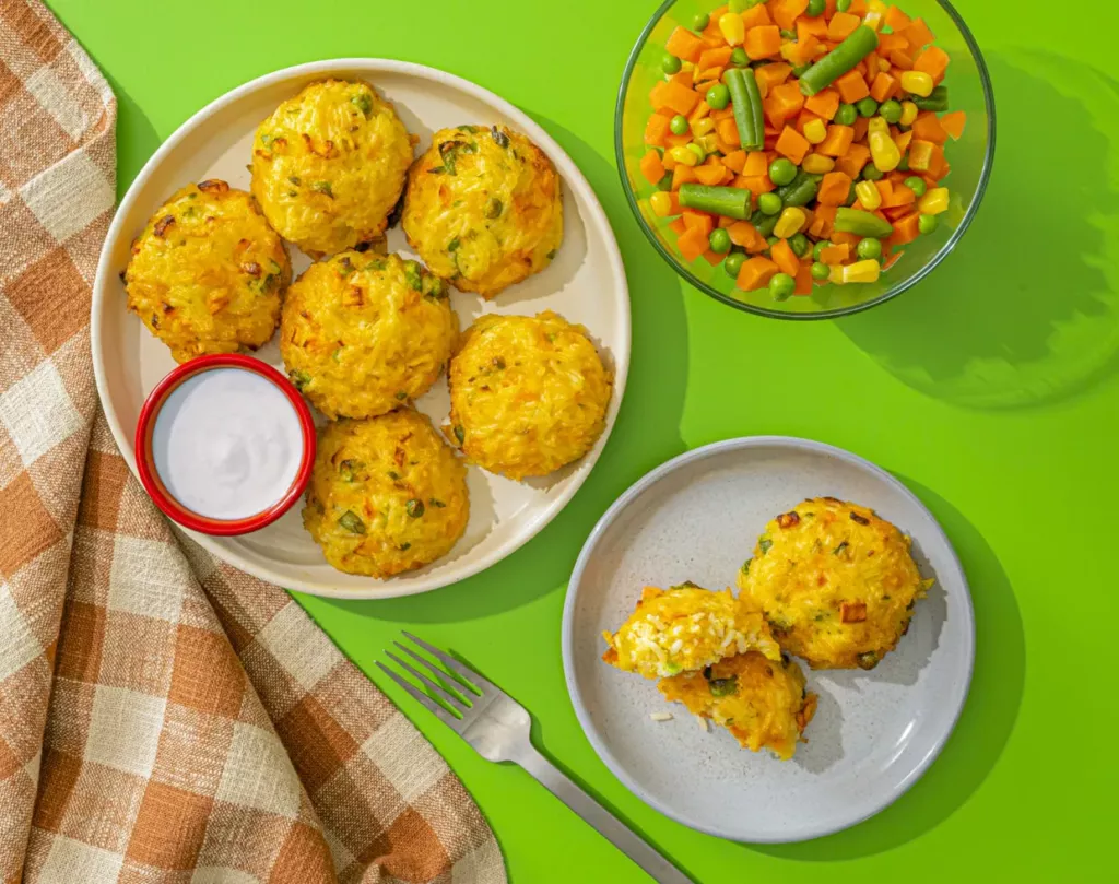 Rice balls on a white plate with a side of mixed diced vegetables, next to an orange and tan checkered napkin on a green background